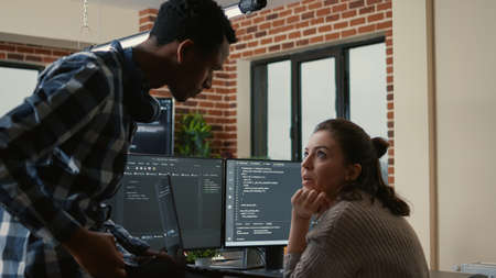 Junior developer sitting down on desk showing laptop with source code to senior dev asking for...
