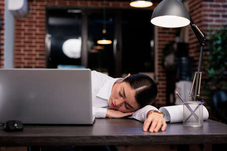 Fatigued business company employee sleeping on desk because of overtime working hours at night. Exhausted agency worker falling asleep in office workspace while working on project.の写真素材