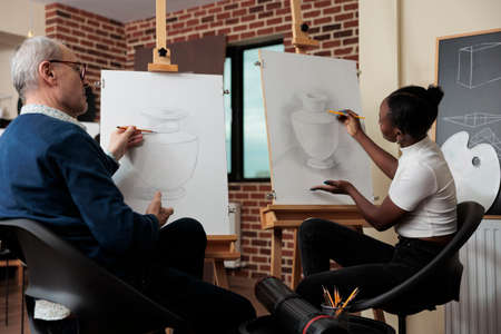 Senior man teacher supervising young student during drawing class in creativity studio developing...