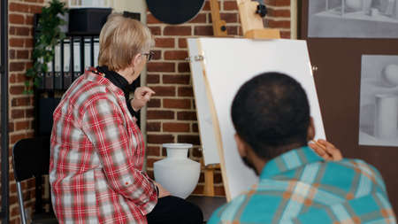 Portrait of senior artist teaching drawing technique at workshop, using art school program to explain artistic hobby. Educational class to draw creative sketch on canvas. Handheld shotの写真素材