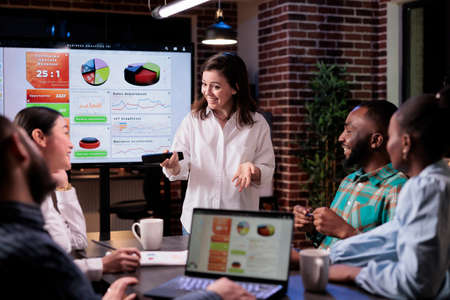 Smiling caucasian woman in late night meeting presenting business analytics to happy team in modern...