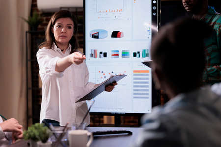 Caucasian business woman holding clipboard pointing at coworker asking question about sales performance in late night meeting. Startup owner in front of wall screen tv with analytics talking to team.の写真素材