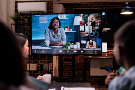 Selective focus on tv screen with african american business owner and clients in online video conference in startup office. Team in late night meeting working overtime brainstorming in internet call.の写真素材