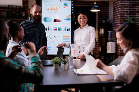 Startup business people talking and smiling at the end of late night sales meeting in corporate office. Marketing team casually discussing marketing strategy in front of wall screen tv with charts.の写真素材