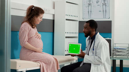 Male doctor holding horizontal greenscreen on tablet display at checkup with future mother....