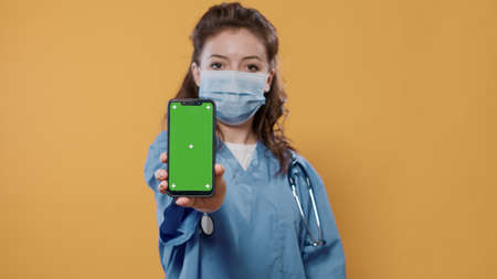 Portrait of medic in hospital uniform wearing surgical mask holding smartphone and texting showing green screen mockup at camera. Doctor with stethoscope and covid protection presenting copy space.の写真素材