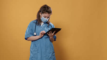 Portrait of woman doctor holding tablet typing patient data wearing hospital uniform and covid protection mask in studio. Medic with stethoscope using digital touchscreen modern technology in scrubs.の写真素材