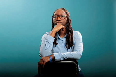 Serious confident woman sitting on chair while looking at camera on blue background. Pretty...