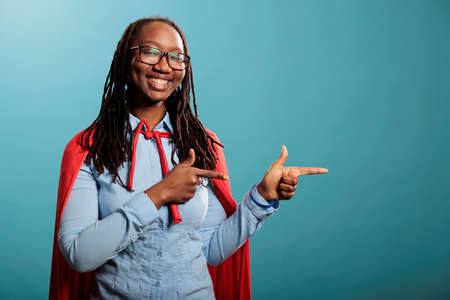 Studio shot of cheerful and optimistic justice defender woman wearing red cape while pointing fingers to right on blue background. Selfless and brave young adult superhero person posing at camera.の写真素材