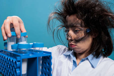 Crazy scientist with dirty face and messy hair analyzing plastic test flasks after laboratory chemical explosion. Mad chemist having rack with test tubes while trying to identify right compound.の写真素材