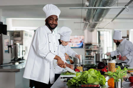 African american gastronomy expert smiling with confidence while cutting fresh vegetables for dinner dish and posing for camera. Food industry workers preparing ingredients for gourmet recipe.の写真素材