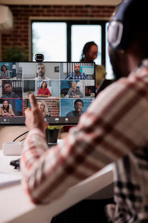 African american with wireless headphones working from home gesturing in video call with colleagues at desk in living room. Startup employee talking with team in internet conference on personal computer.の写真素材