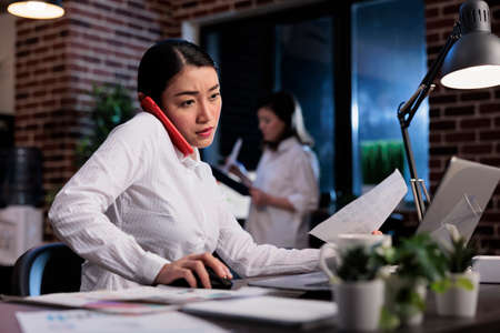 Asian businesswoman talking on cellphone with agency colleague about startup project. Young adult entrepreneur sitting in office workspace while talking on smartphone device with executive manager.の写真素材