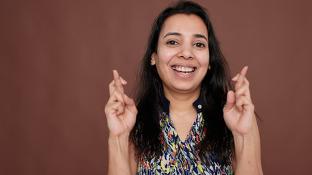 Indian woman holding fingers crossed for good luck in studio, feeling positive about success belief. Hopeful model waiting for miracle, wanting to fulfill wish with fortune, smiling.の写真素材