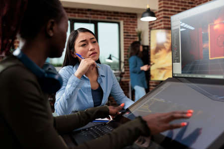 Multiethnic creative game creators sitting at desk with multiple displays while working with 3D assets. Digital interactive entertainment industry workers working with CGI.の写真素材