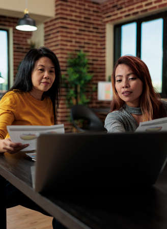 Businesswomen holding business papers discussing management strategy while analyzing graphs on...