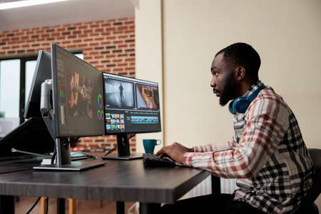 Video production house employee sitting at multi monitor workstation while editing movie frames using advanced software. African american graphic footage editor enhancing project quality.の写真素材