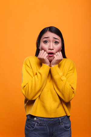 Frightened scared young adult woman covering mouth with hands in fear on orange background. Attractive brunette asian lady left stunned speechless while hearing tragic news on television.の写真素材