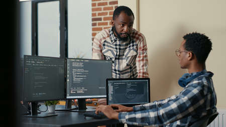 Software developer sitting at desk with multiple screens and laptop running code talking with...