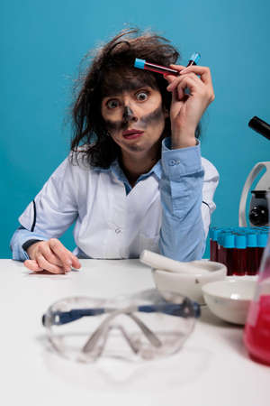 Potrait of crazy looking mad silly chemist with messy hair and dirty face having glass test tubes filled with blood samples after laboratory explosion. Maniac scientist sitting at desk. Studio shotの写真素材