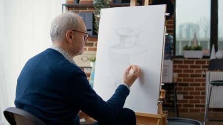 Old man drawing vase model on canvas at art class lesson, attending workshop with young students and senior teacher. Elder person learning to draw sketch to develop skills and personal growth.の写真素材