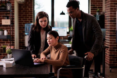 Diverse team of colleagues working with paralyzed employee sitting in wheelchair, suffering from chronic disability and impairment. Business people using laptop, health condition.の写真素材