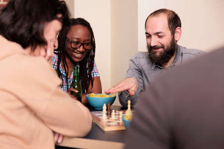 Laughing close friends sitting at table in living room while playing chess. Multiethnic intelligent...