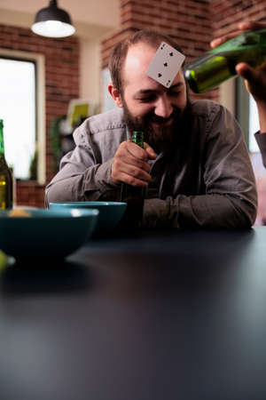 Cheerful man with plastic card on forehead laughing with friends while hanging out together. Young adult person enjoying fun leisure activity at home in living room while having snacks and beverages.の写真素材