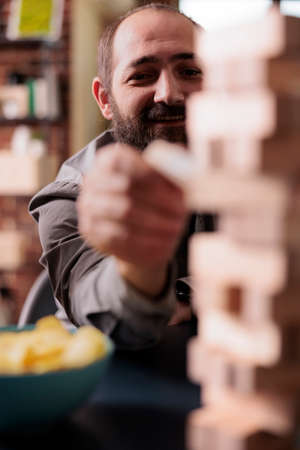 Selective focus of smiling man pulling wooden cube from blocks tower structure. Happy person...