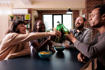 Happy young diverse people sitting at table while relaxing with beer and snacks. Positive multiethnic group of friends enjoying drinking and having fun together at home in living room.の写真素材