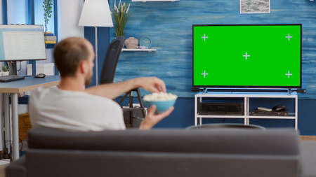 Man relaxing on couch looking at green screen on tv watching movie while eating popcorn in modern living room. Person having a good time sitting on sofa looking at television with chroma key display.の写真素材