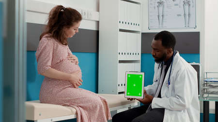 Obstetrician holding greenscreen on digital tablet at checkup visit with expectant patient. Medic using isolated copyspace with mockup template and blank chroma key background. Tripod shot.の写真素材