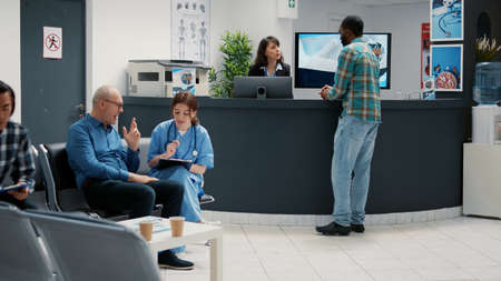 Reception desk with diverse patients waiting in lobby for checkup visit, people writing report at counter and mother with child in waiting area. Appointments with physician and medical staff.の写真素材