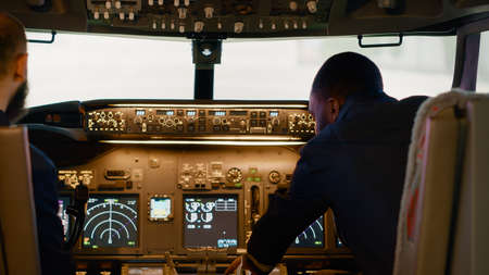 Diverse team of captain and copilot in cockpit flying airplane, using control panel buttons to...