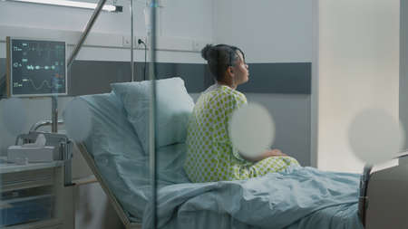 Portrait of young adult with sickness sitting in hospital ward bed to receive medical care and treatment. Patient waiting on medic to help with pain and disease at healthcare clinic.の写真素材