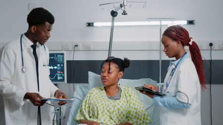 Team of african american doctors consulting sick patient in bed. Medical specialists discussing about healthcare treatment and medicine to cure illness while woman holding tablet.の写真素材