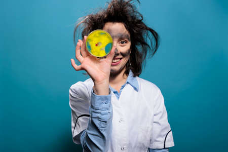 Crazy scientist with dirty face and messy hair with petri dish glass plate having dangerous bacterial sample pointing towards camera. Lunatic goofy looking biochemistry expert grinning creepy.の写真素材