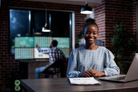 Portrait of professional broker sitting at desk while coworker in background working at multi monitor workstation. African american employees in hedge fund company office.の写真素材