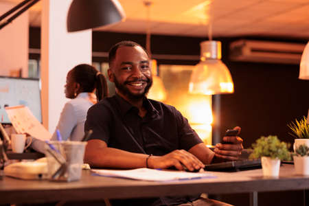 Portrait of male employee using smartphone app at office job, working with mobile phone and computer...