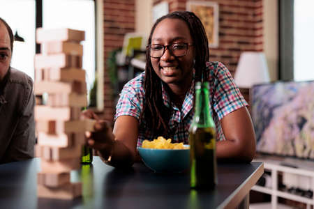 Happy african american woman pulling wood block from tower structure game. Confident joyful person playing society games with multicultural group of friends while sitting at home in living room.の写真素材
