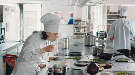 Woman working as cook testing bad soup for gourmet dish, making mistake in preparing culinary recipe. Authentic chef doing taste test with sauce, cooking in professional gastronomy kitchen.の写真素材