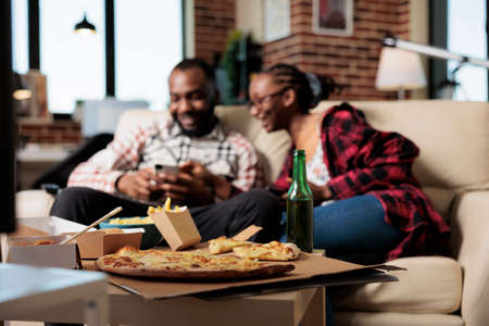 African american partners with takeaway food using mobile phones to browse internet, watching film on television and eating fast food delivery. Leisure activity with takeout and tv.の写真素材
