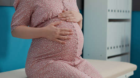 Pregnant patient attending checkup visit with obstetrician, sitting on bed in medical office. Woman...