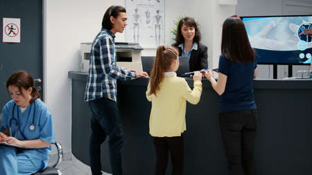 Mother and little girl writing checkup report at hospital reception desk to attend medical appointment. Patients having healthcare examination to receive support and assistance.の写真素材