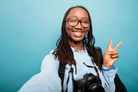 Positive happy photography entusiast with DSLR camera making peace sign with fingers while taking selfie photo. Confident optimistic young woman standing on blue background while gesturing victory.の写真素材