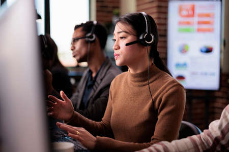 Asian female receptionist working at customer service helpdesk, talking to clients on helpline support at call center. Sales consultant using remote telecommunication assistance in office.の写真素材