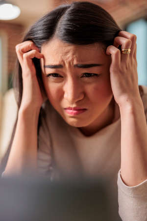 Young asian woman having migraine and headache while trying to work from home. Tired female on couch covering forehead and exhausted red eyes unhappy sick upset ill fever depresionの写真素材