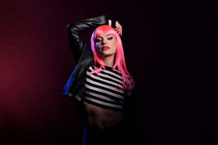 Confident young adult posing in dark pink shadow over background, wearing rocker leather jacket to...