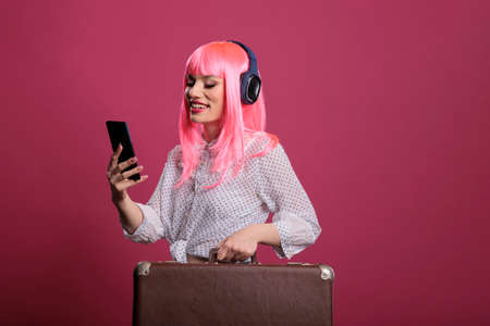 Female model using smartphone and carrying suitcase luggage in front of studio camera. Travelling on...