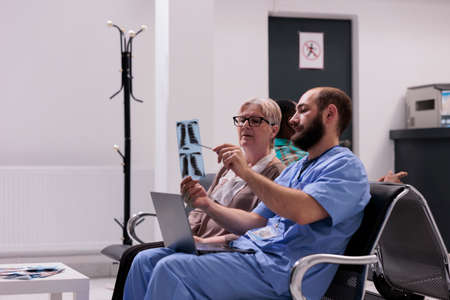 Medical assistant showing x ray to elderly patient, sitting in hospital reception lobby. Nurse explaining radiography scan results to woman with disease, talking about treatment and diagnosis.の写真素材